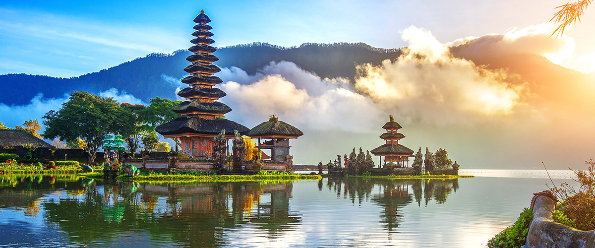 Im Vordergrund trägt eine Person in traditioneller balinesischer Kleidung einen Opferkorb. Dahinter liegt der Wassertempel Pura Ulun Danu Bratan am See, umgeben von Bergen.
