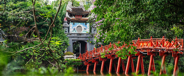 Die berühmte rote Huc-Brücke aus Holz führt in Hanoi über den Hoan-Kiem-See. Man sieht die Brücke, Wasser, Bäume und den Eingang zum Jadebergtempel.