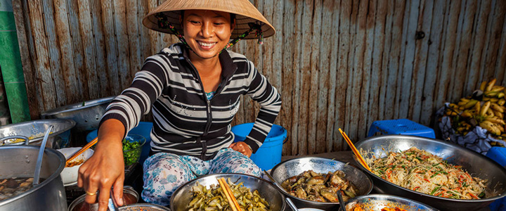 Streetfood in Vietnam: Eine Verkäuferin sitzt vor Schüsseln mit Suppen, gebratenem Gemüse und Nudeln, einen typischen spitzen Strohhut auf dem Kopf.